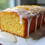 A close-up of a moist Greek Yogurt Lemon Loaf Cake with thick white glaze dripping down the sides and lemon zest on top.