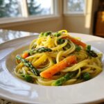 A close-up of a white bowl filled with bright Pasta Primavera featuring spaghetti, asparagus, carrots, and peas.