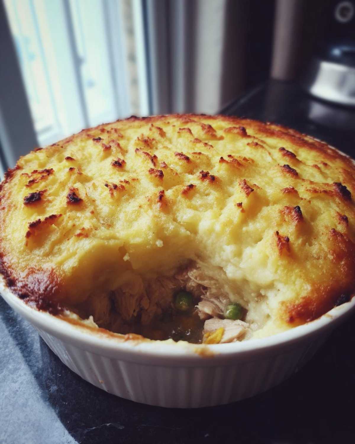 A close-up of a baked Rotisserie Chicken Shepherd's Pie with a golden, textured mashed potato topping, showing the chicken and peas filling underneath.