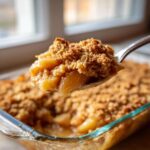 A spoonful of warm, gooey Apple Crisp with a crunchy oat topping being lifted from a glass baking dish.