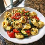 A close-up of a white bowl filled with Tortellini with Summer Veggies, featuring roasted tomatoes, zucchini, and fresh basil.