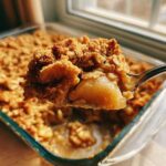A spoonful of warm, gooey Apple Crisp being lifted from a glass baking dish.