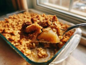 A spoonful of warm, gooey Apple Crisp being lifted from a glass baking dish.