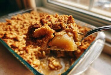 A spoonful of warm, gooey Apple Crisp being lifted from a glass baking dish.